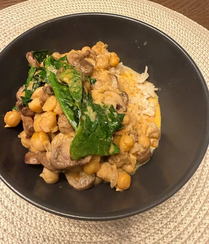 Curry de champiñones y garbanzos con espinacas en plato blanco sobre mantel en mesa de madera oscura.